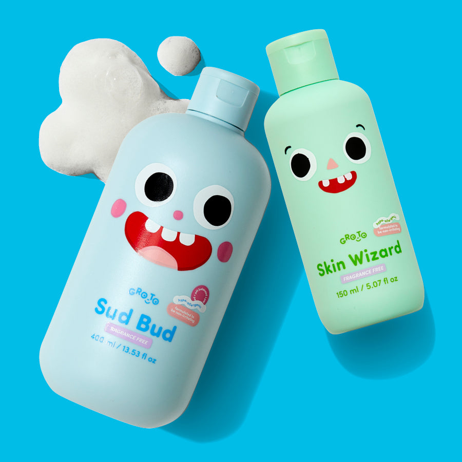Two colorful bottles with cartoon faces, labeled Sud Bud and Skin Wizard, are placed on a bright blue background. White foam is next to the blue Sud Bud bottle, suggesting a playful bath or skincare theme.