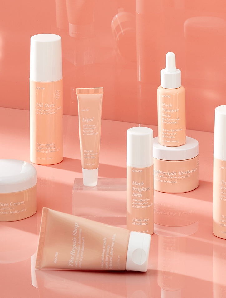A collection of peach-colored skincare and beauty products, including creams, serums, and tubes, displayed neatly on a glossy peach surface with reflections. The products have minimalist labels and white caps.