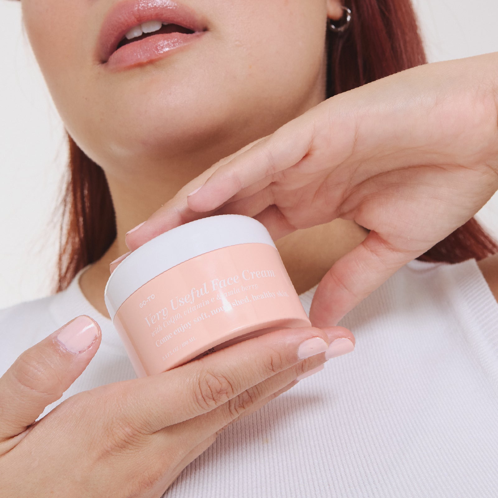 A person wearing a white sleeveless top holds a pink jar of Very Useful Face Cream near their chin. Their lips are glossy, and their nails are painted a soft pastel pink.