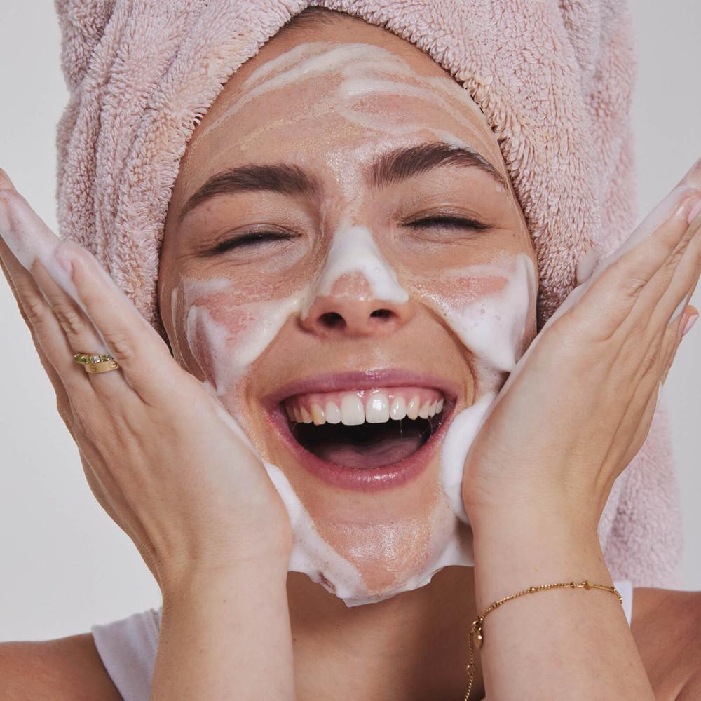 A smiling woman with a pink towel wrapped around her head applies foamy facial cleanser to her face, holding her hands to her cheeks. She appears happy and is looking directly at the camera.