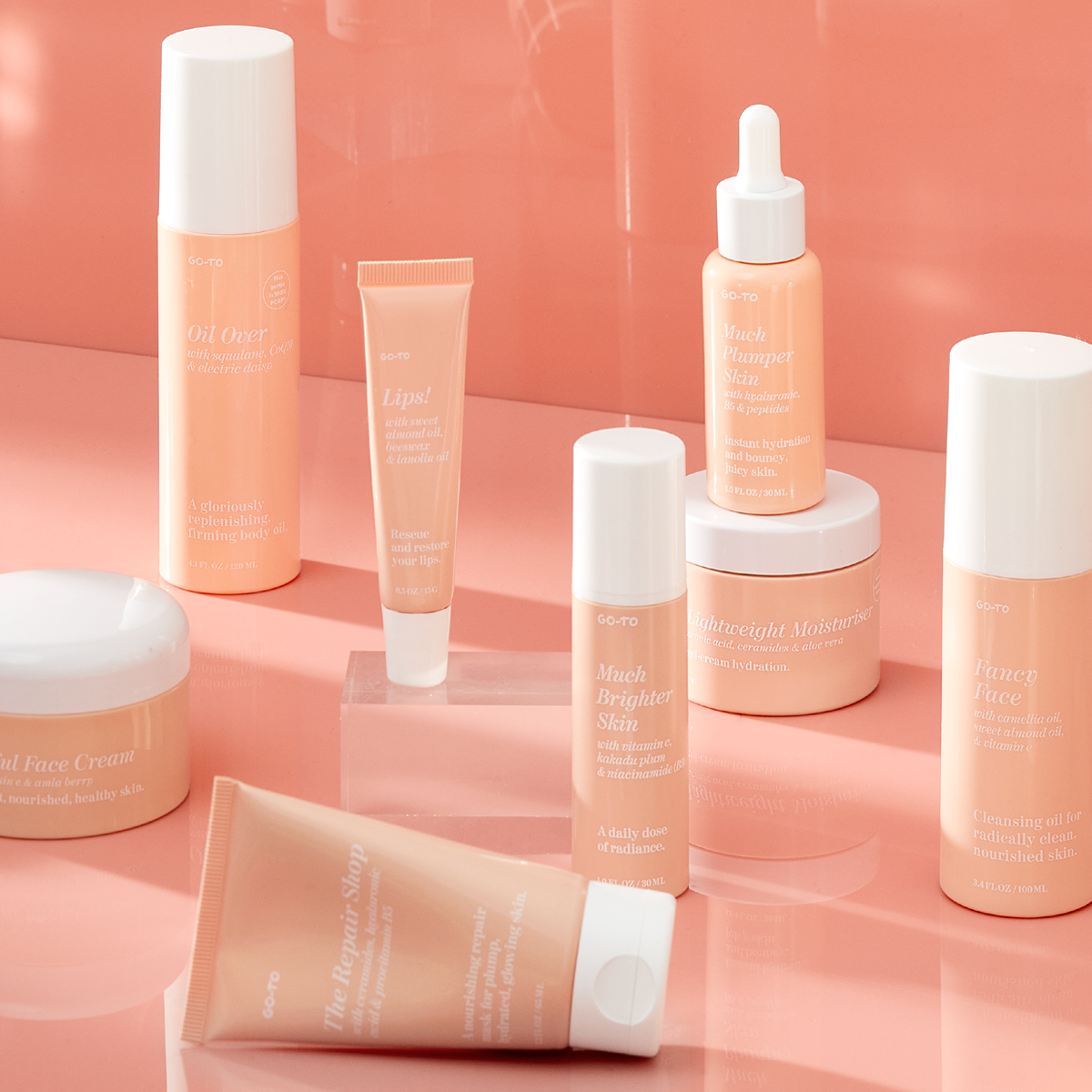 A collection of peach and white skincare products, including creams, serums, oils, and a lip balm, arranged neatly on a glossy peach-colored surface. All containers feature minimalist labels.