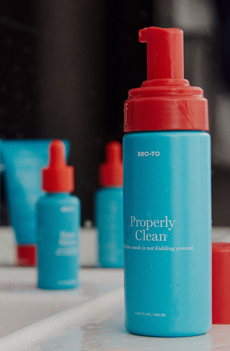 A blue bottle with a red pump labeled BRO-TO Properly Clean sits on a white surface, reflected in a mirror. Another blue skincare product is blurred in the background.