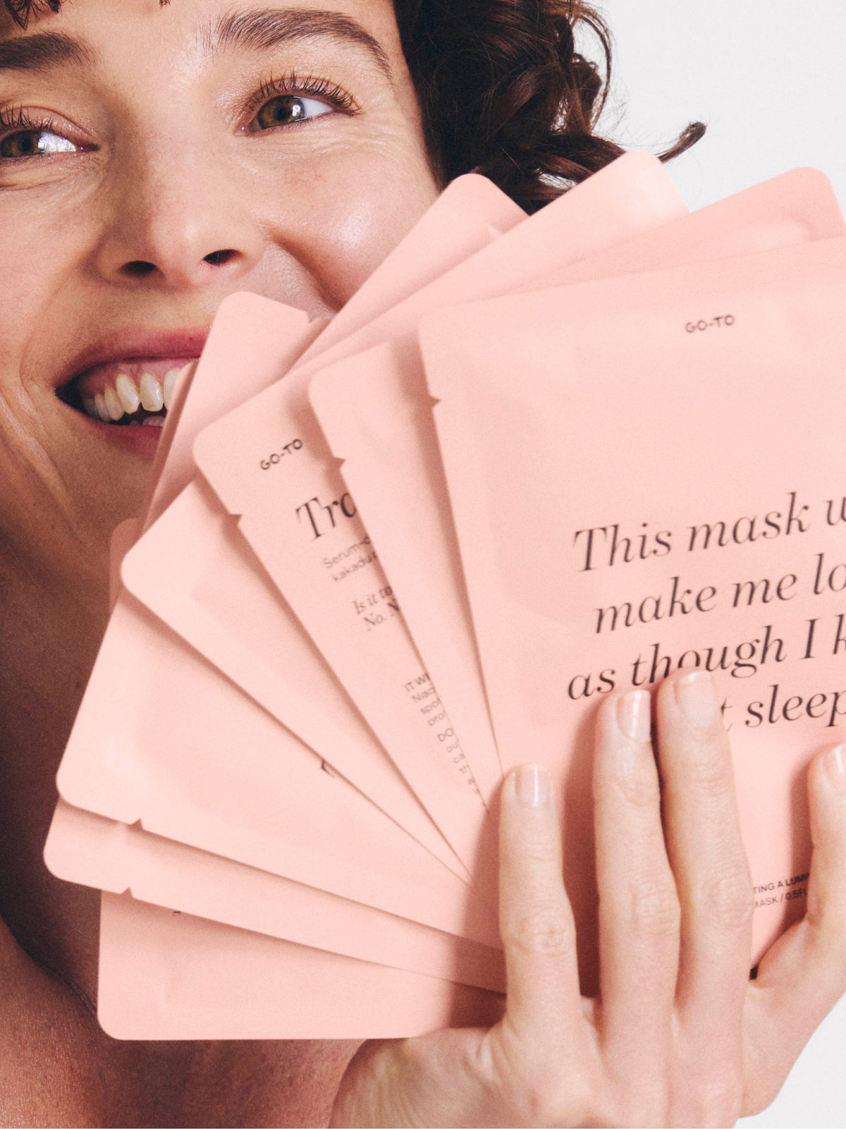 Smiling woman holding a fan of pink Go To Skincare sheet mask packets with printed text visible on the packaging