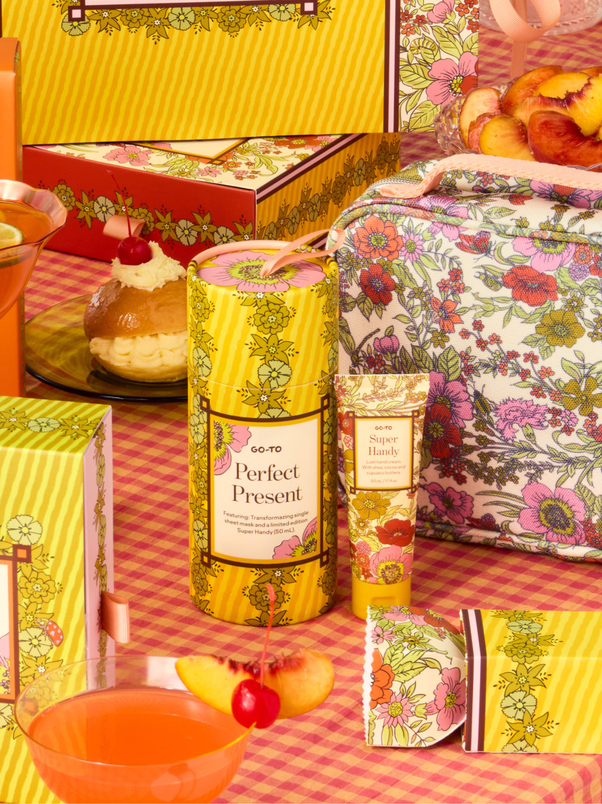 Go-To Skincare Perfect Present gift set with floral packaging and pouch alongside a yellow cylindrical package and Super Handy hand cream on a colorful picnic table setup