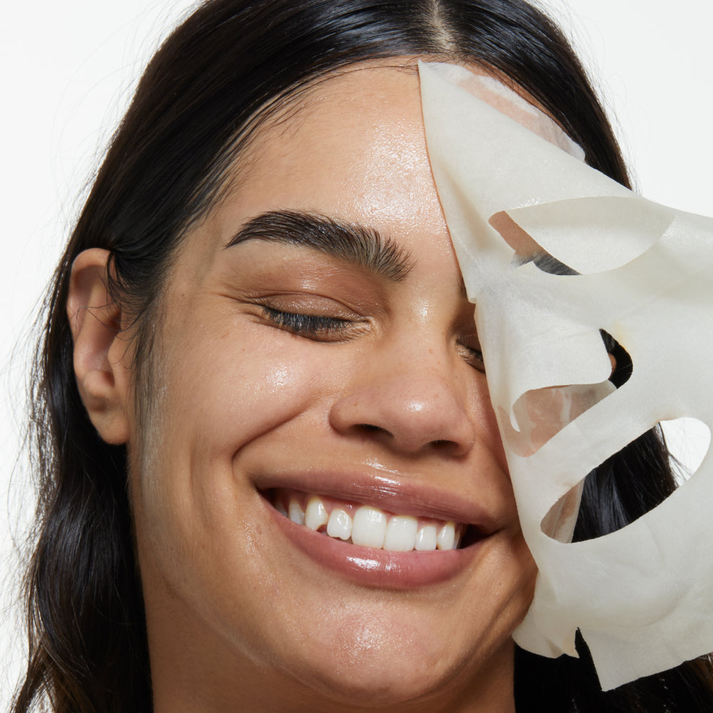 A woman with smooth, glowing skin smiles with her eyes closed while peeling off a white sheet mask from her face.