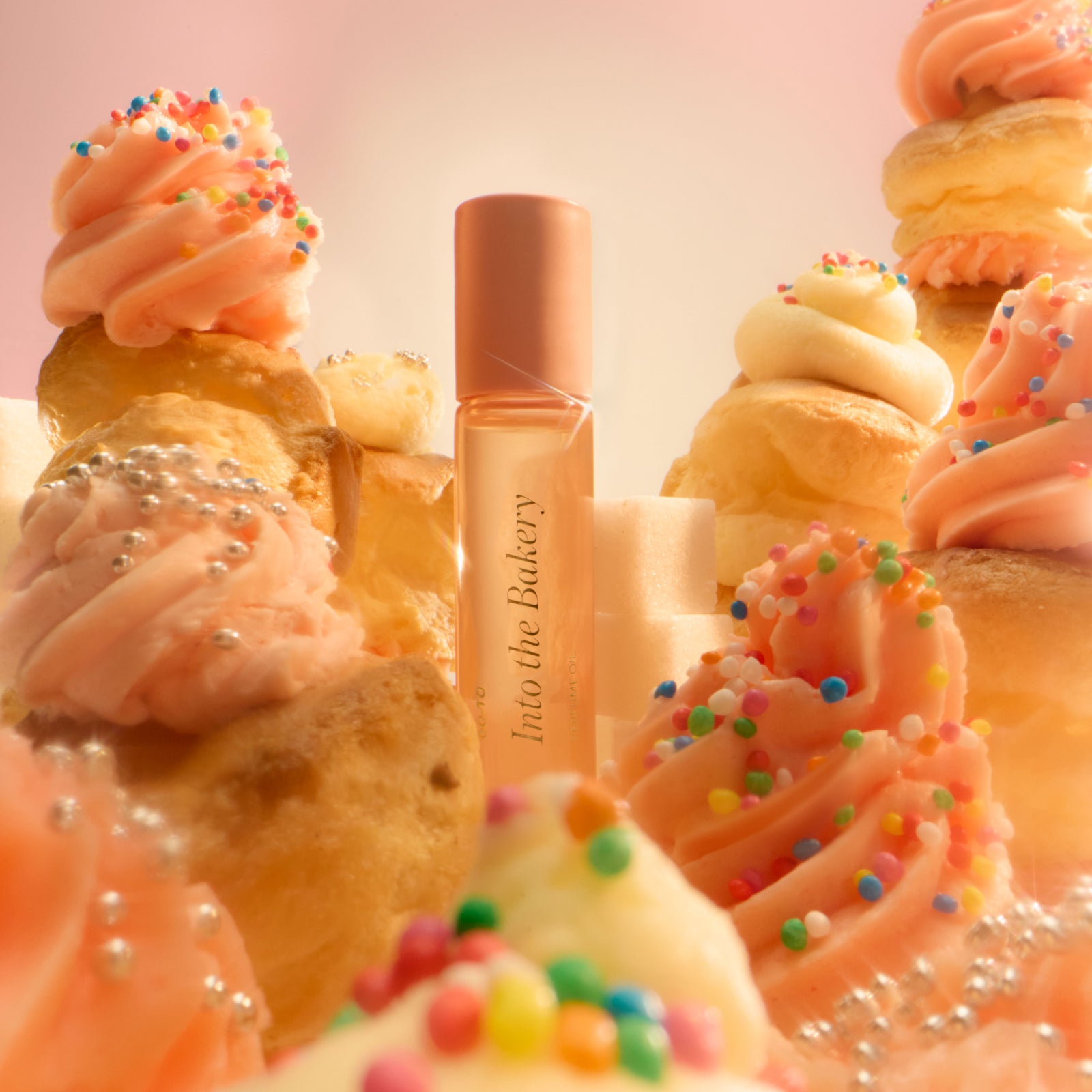 A perfume bottle labeled Into the Bakery stands surrounded by pink frosted cupcakes, cream puffs, and pastries decorated with colorful sprinkles and silver pearls, all in soft, warm lighting.