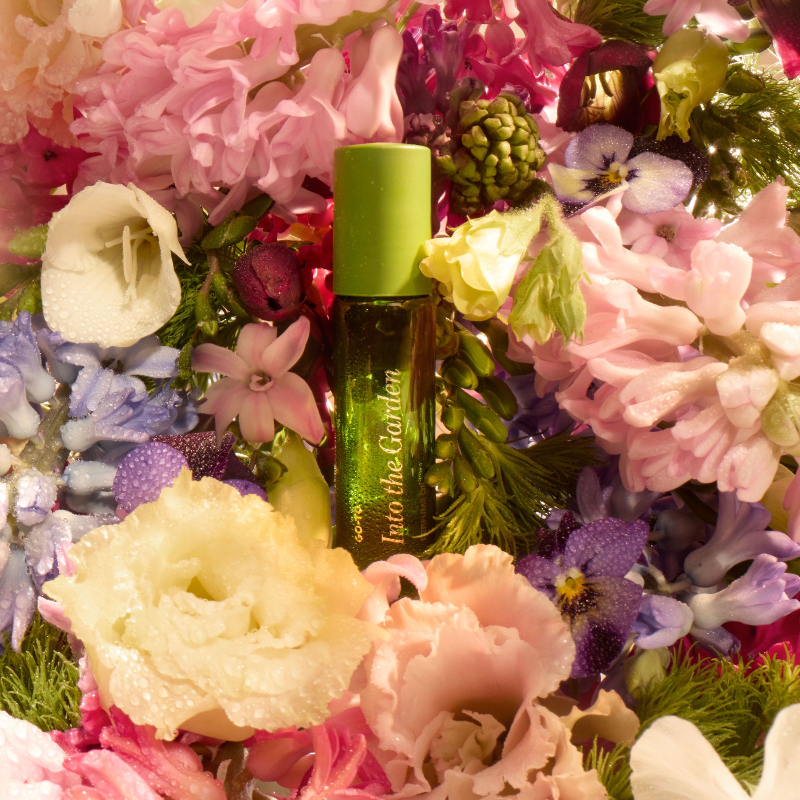 A green perfume bottle labeled Into the Garden is surrounded by an abundance of colorful, dewy flowers in shades of pink, purple, white, and yellow.