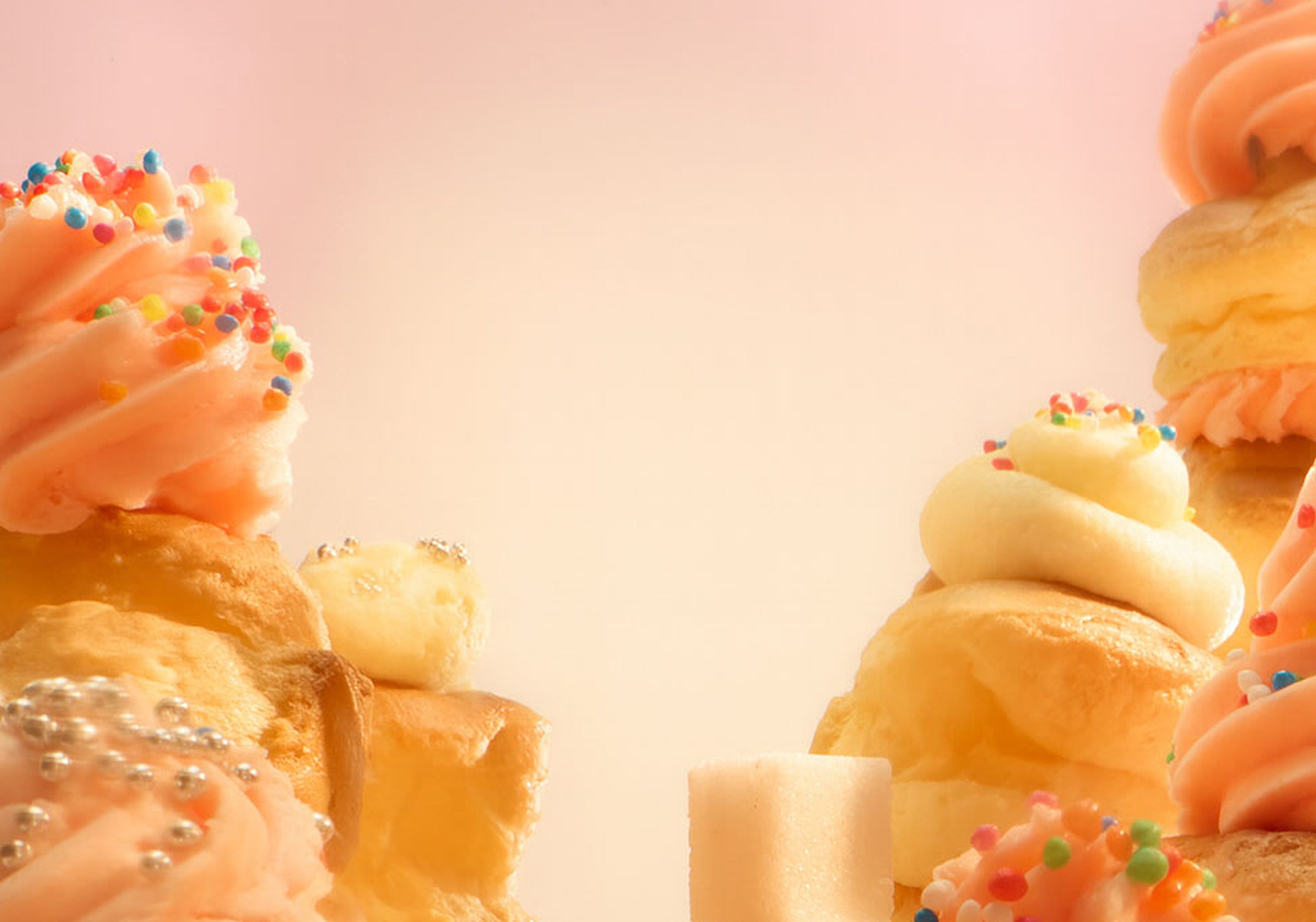 Close-up of pastel pink and white frosted pastries topped with colorful sprinkles, set against a soft, light pink background. The arrangement leaves a bright empty space in the center.