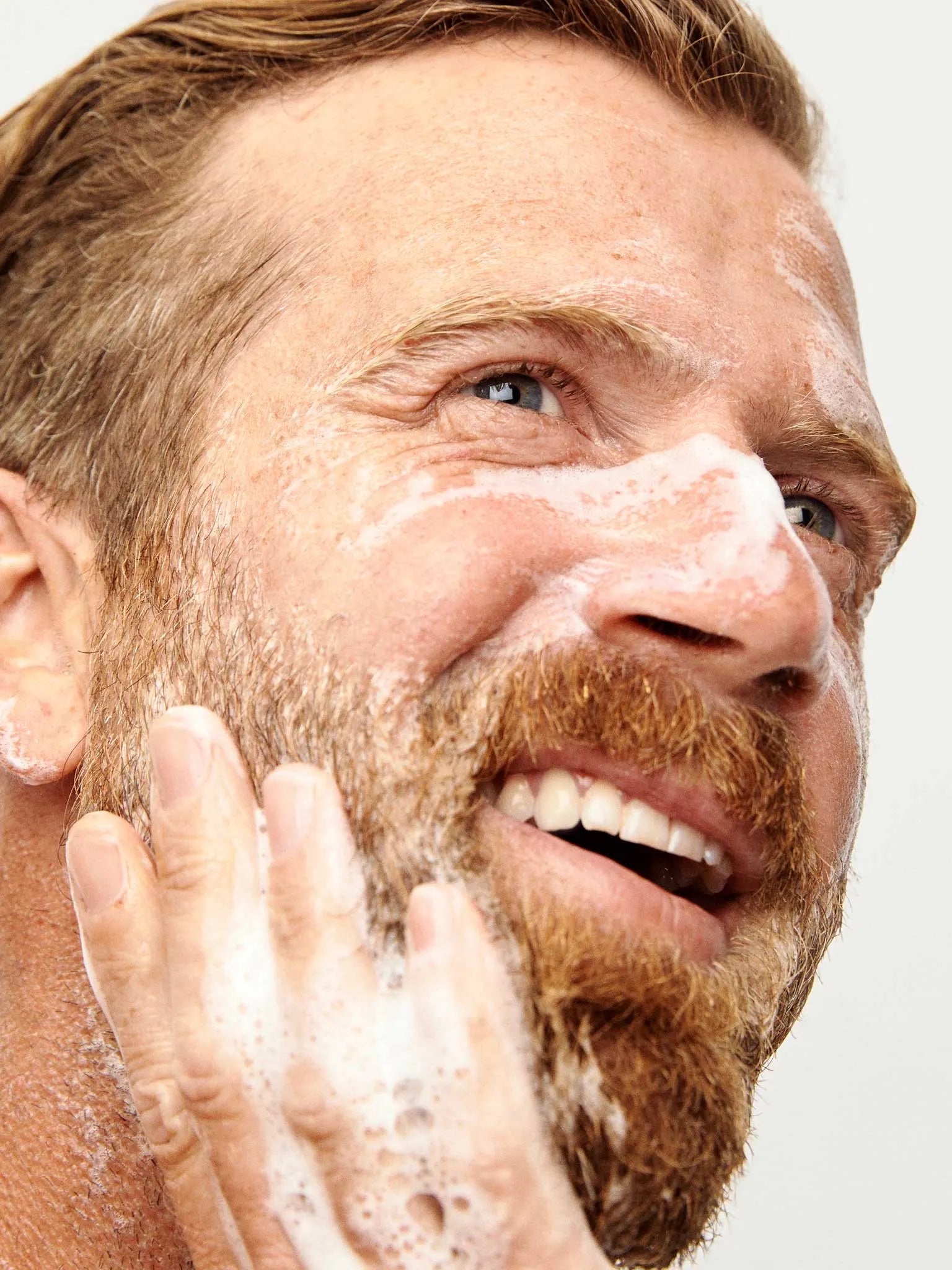 A man with a beard smiles while washing his face, spreading foamy cleanser on his skin with his hand.