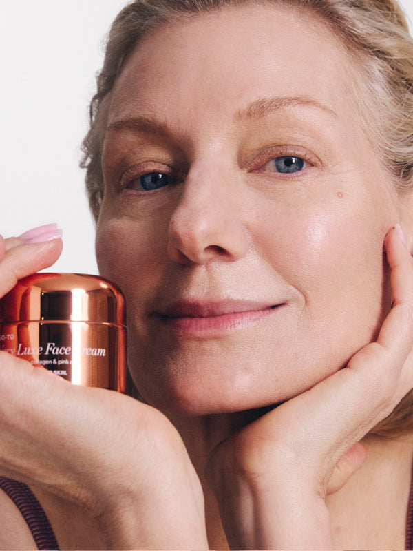 Woman smiling, holding a jar of Luxe Face Cream close to her face, showcasing smooth skin and a sense of satisfaction.