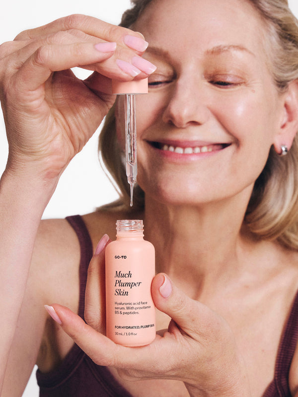 Smiling woman applies Go-To Much Plumper Skin serum with a dropper, highlighting skincare routine and product packaging.